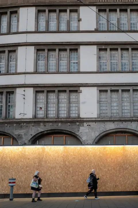 Deux passantes longent un mur de bois brut devant une façade ancienne, Munich