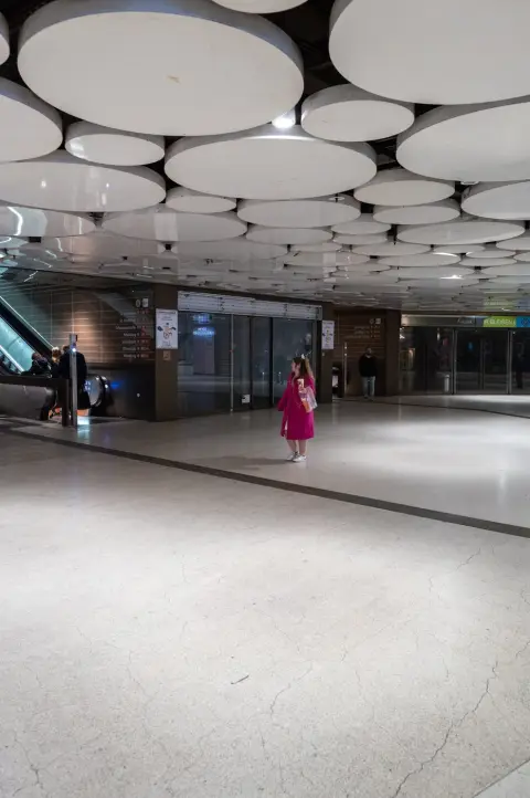 Une passante en manteau rose traverse un hall de gare au plafond ondulé, Munich