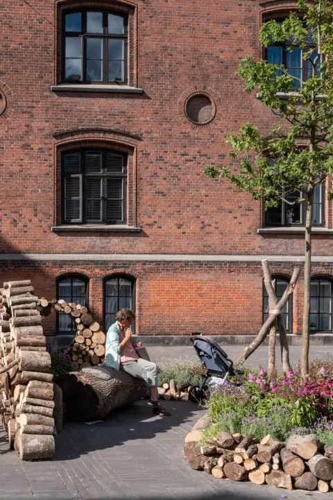 Une femme assise parmi des bûches empilées au pied d’un mur de briques, Odense