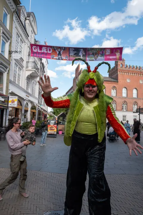 Un échassier costumé en sauterelle verte ouvre les bras, Odense