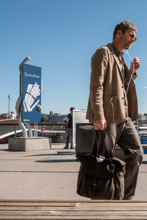 Un homme en veste beige, sacoche à la main, près du panneau de Tjuvholmen, Oslo