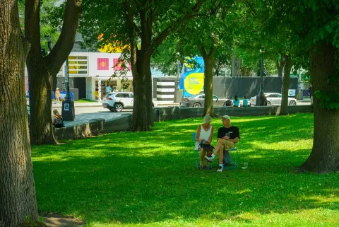 Un couple assis sur des chaises pliantes à l’ombre des arbres, devant les tentes d’un festival, Québec