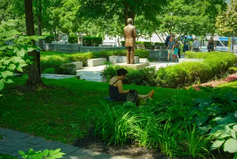 Une femme assise dans l’herbe près d’une statue de bronze, au milieu des massifs, Québec