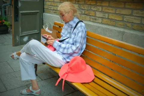 Une dame lit son téléphone sur un banc, son chapeau corail posé à côté d’elle, Québec