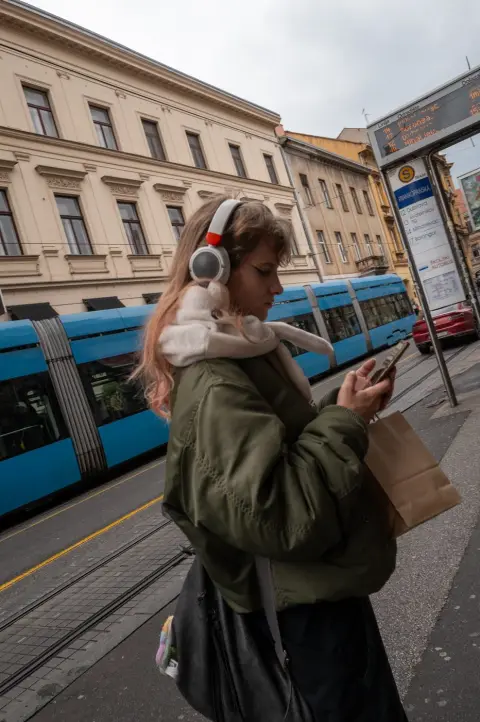 Une passante aux écouteurs et à la peluche sur l'épaule, tramway bleu en fond, Zagreb