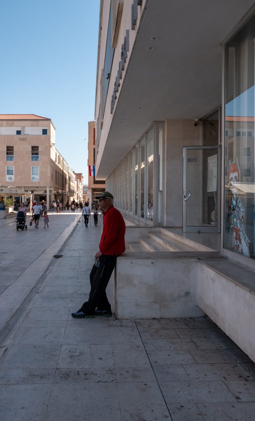 A man in a red jacket leaning against a low wall in the street, Zadar