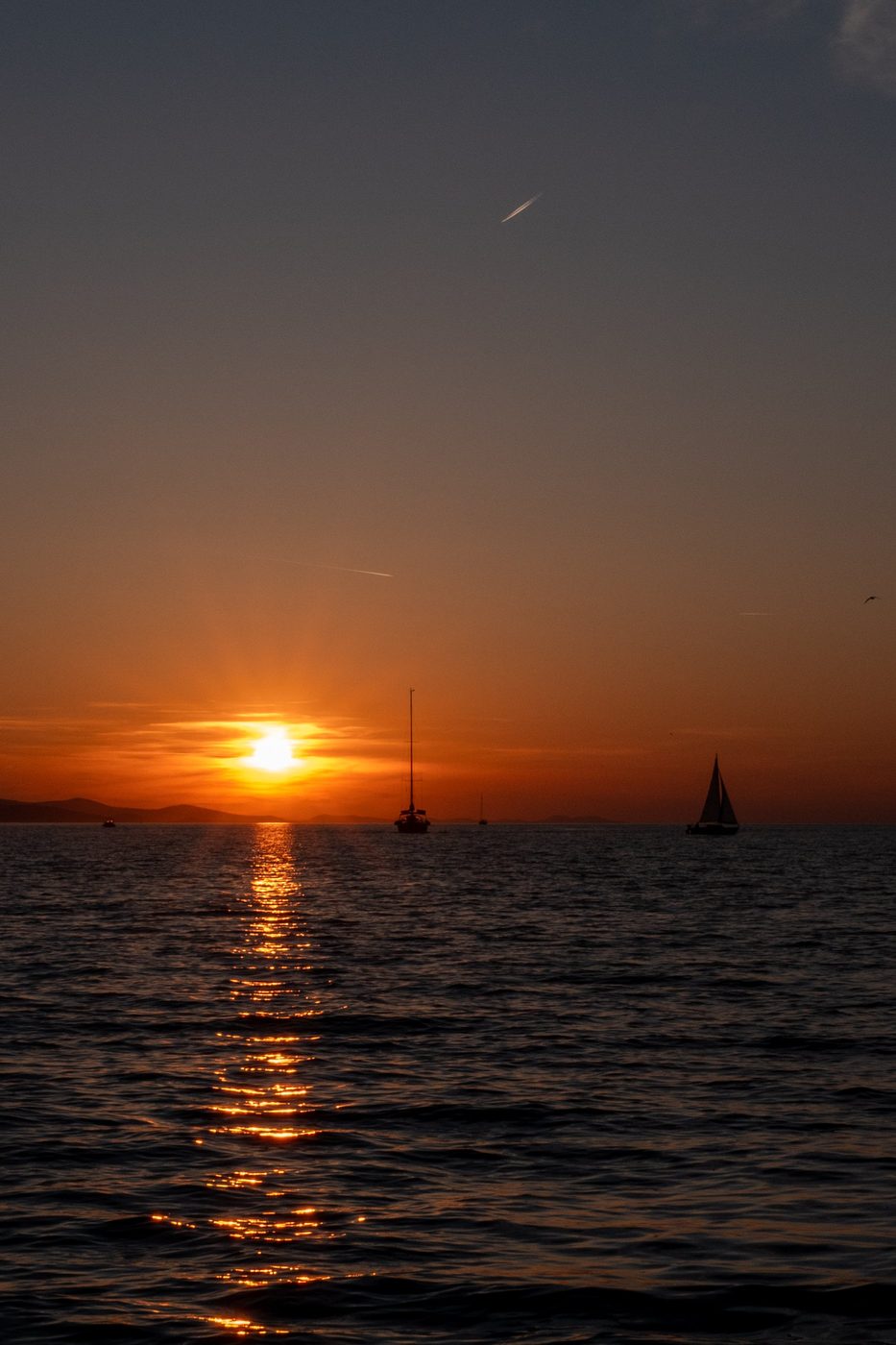 Boats at anchor facing the setting sun, a plane's contrail in the sky, Zadar