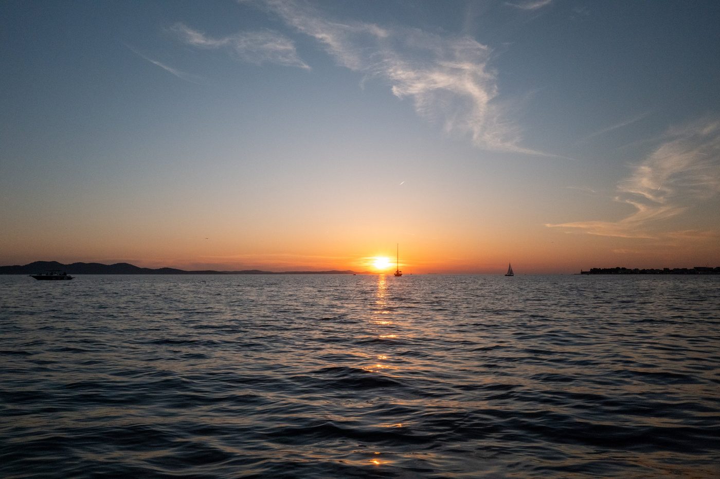 Sailboats anchored offshore beneath a blazing sky, the town's silhouette in the distance, Zadar