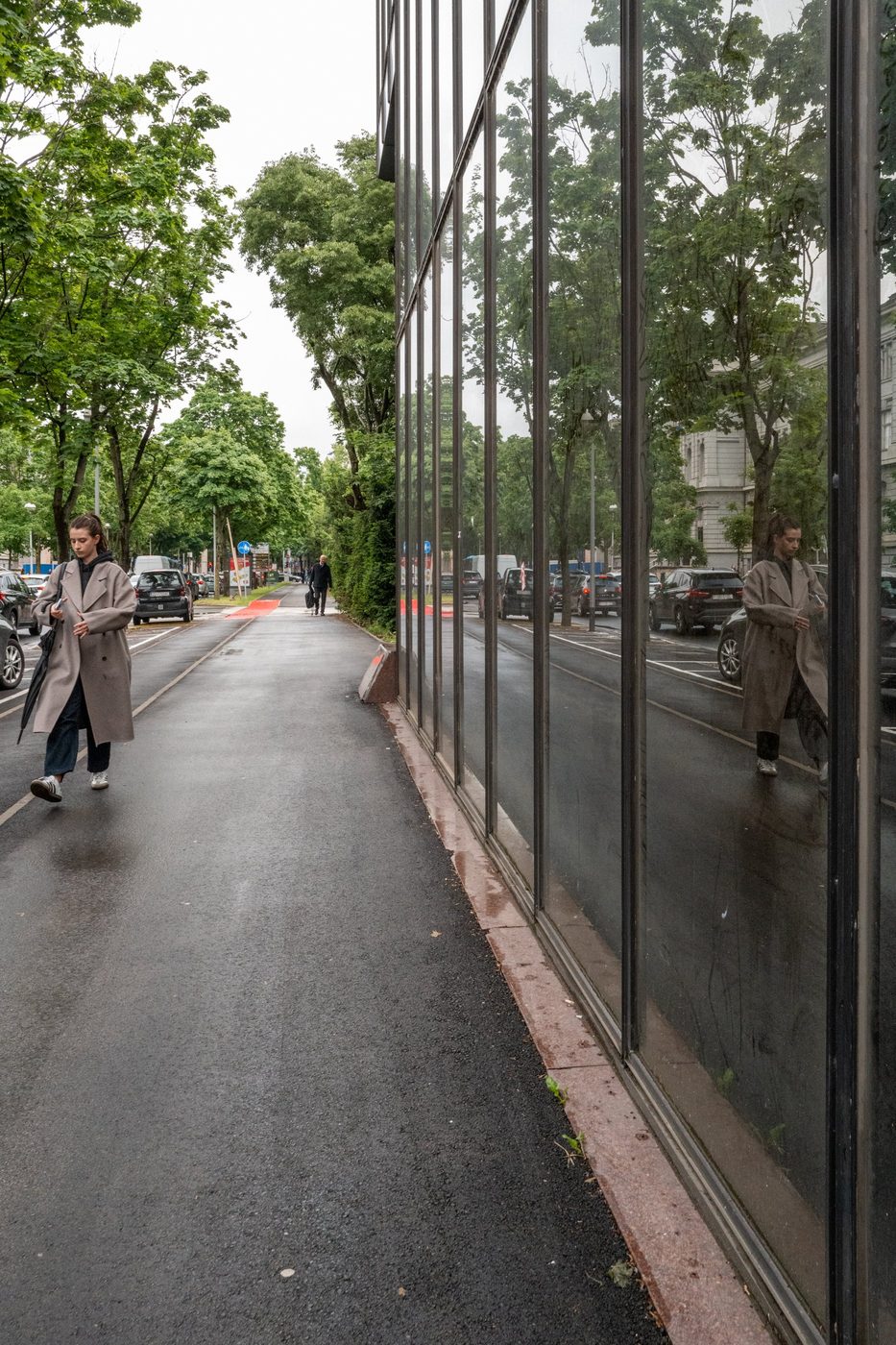 A passerby in a beige trench coat reflected in a glass façade, Zagreb