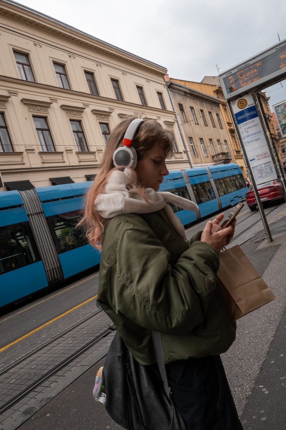 A passerby with earphones and a stuffed toy on her shoulder, blue tram in the background, Zagreb