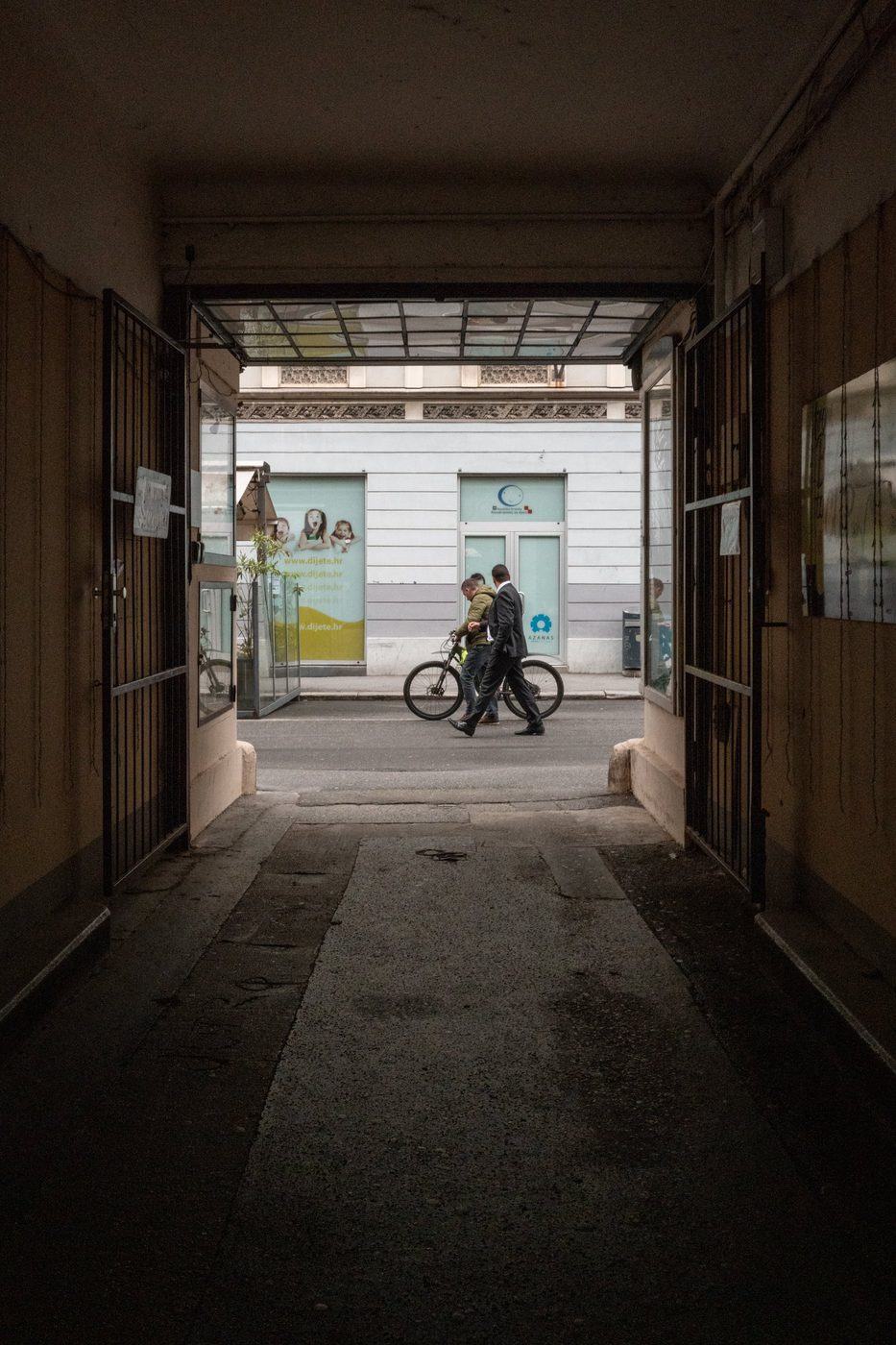 Dark porch opening onto a street where two men and a bicycle pass by, Zagreb