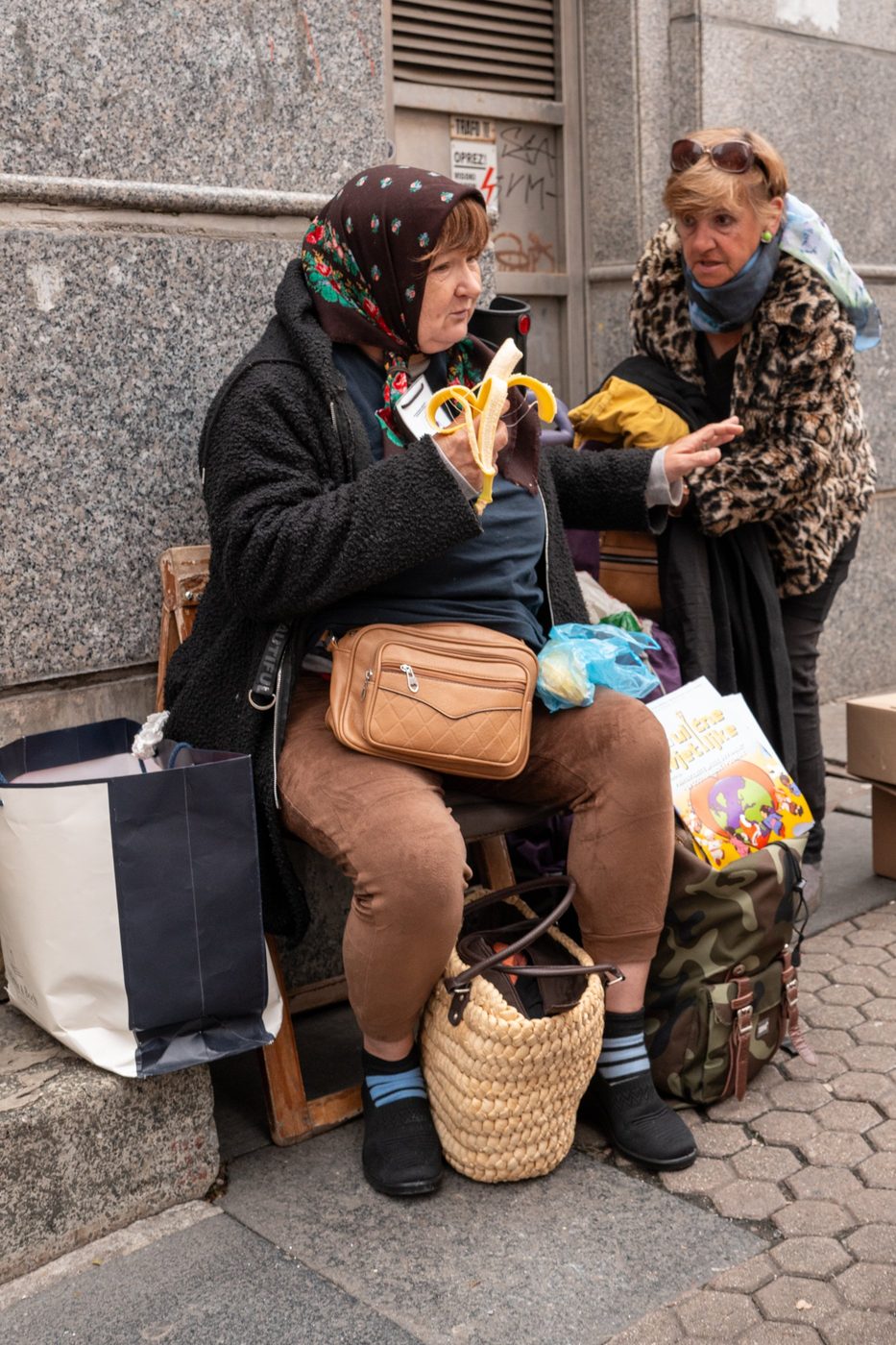 Two elderly women chatting on a bench, one biting into a banana, Zagreb